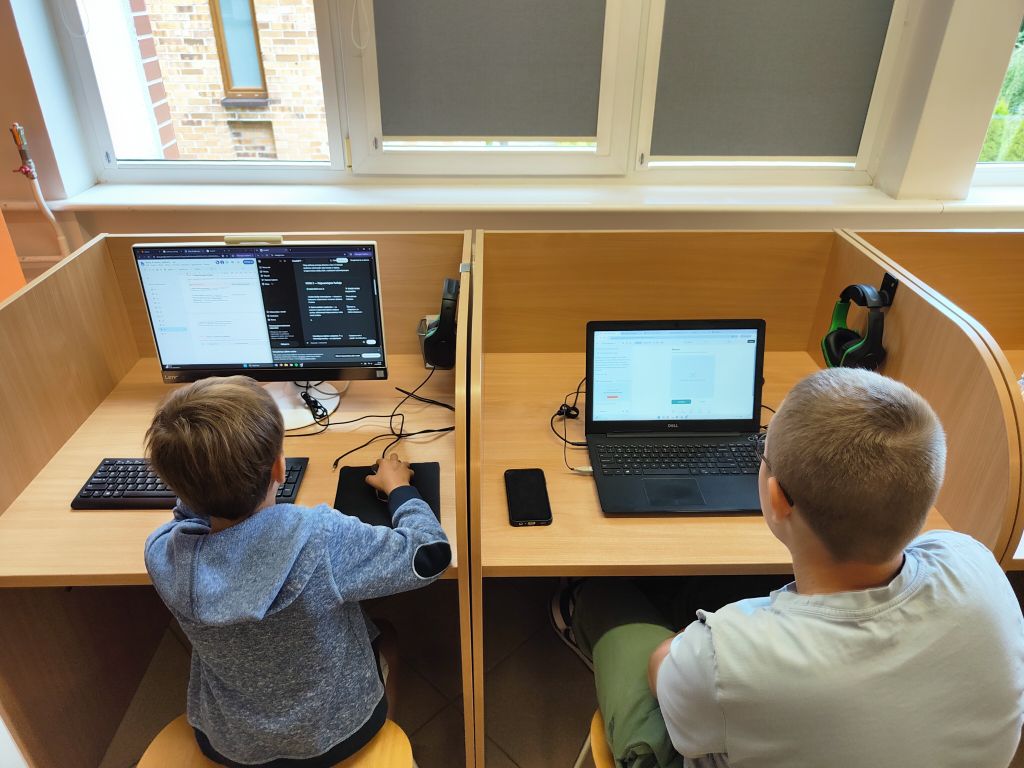Dwóch chłopców siedzi przy oddzielnych stanowiskach komputerowych w boksach bibliotecznych, pracując na monitorze stacjonarnym i laptopie.