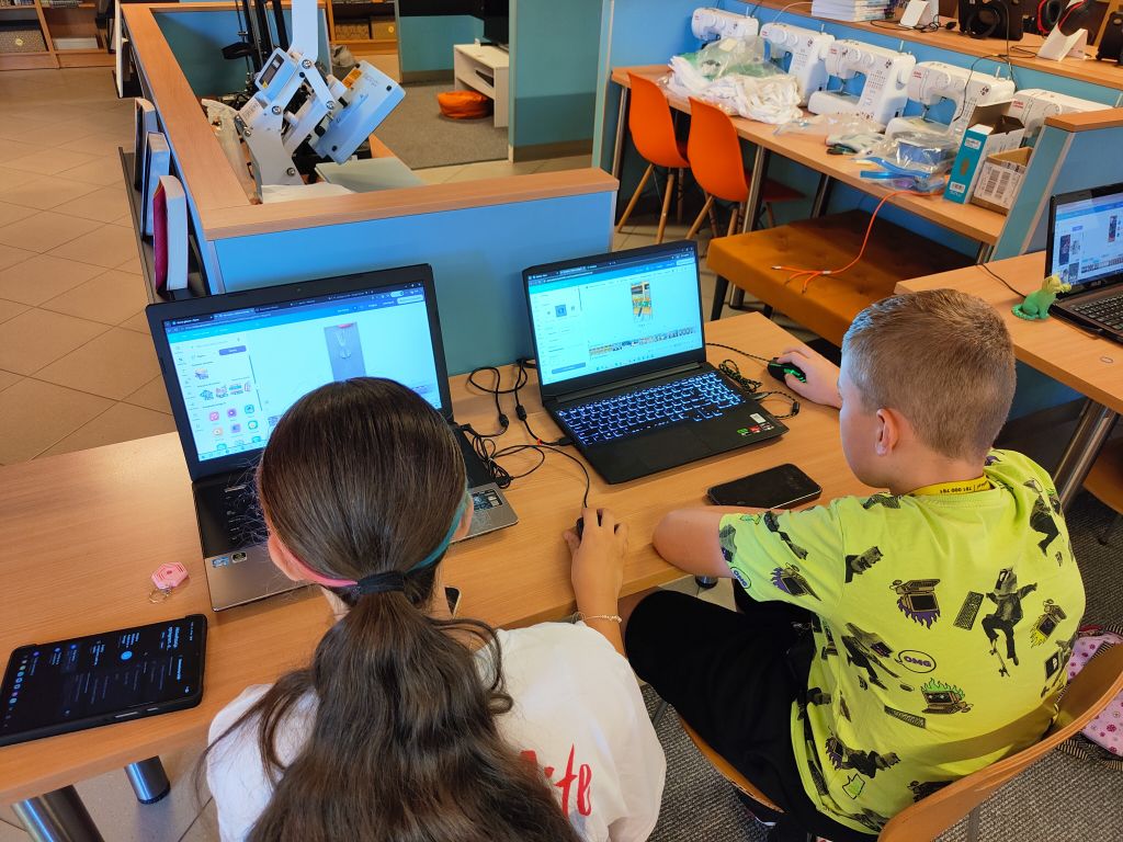 Dwoje dzieci siedzi obok siebie przy biurku i pracuje na dwóch oddzielnych laptopach. Chłopiec w zielonej koszulce używa myszki. W tle widać pracownię krawiecką z maszynami do szycia.