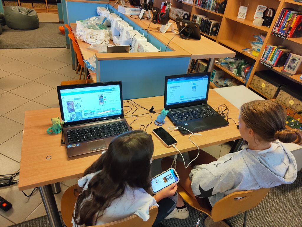 Dwie dziewczynki siedzą przy biurku z laptopami. Dziewczynka na pierwszym planie trzyma smartfon i patrzy na jego ekran. Na stole, obok laptopów, stoją małe, kolorowe figurki kotów.