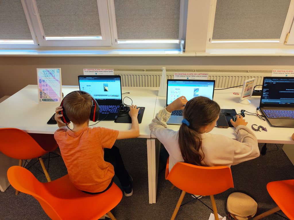 Chłopiec w pomarańczowej koszulce i dziewczynka siedzą tyłem przy białym blacie pod oknem. Oboje mają na uszach duże słuchawki nagłowne i pracują na laptopach. Między nimi stoi tabliczka z informacją.
