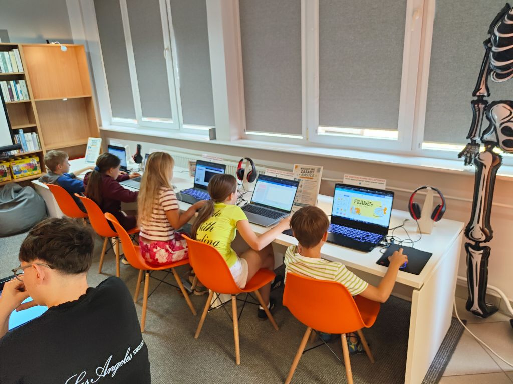 Dzieci siedzące na pomarańczowych krzesłach przy długim białym stole tworzą projekty na laptopach. W tle widoczny jest model szkieletu.
