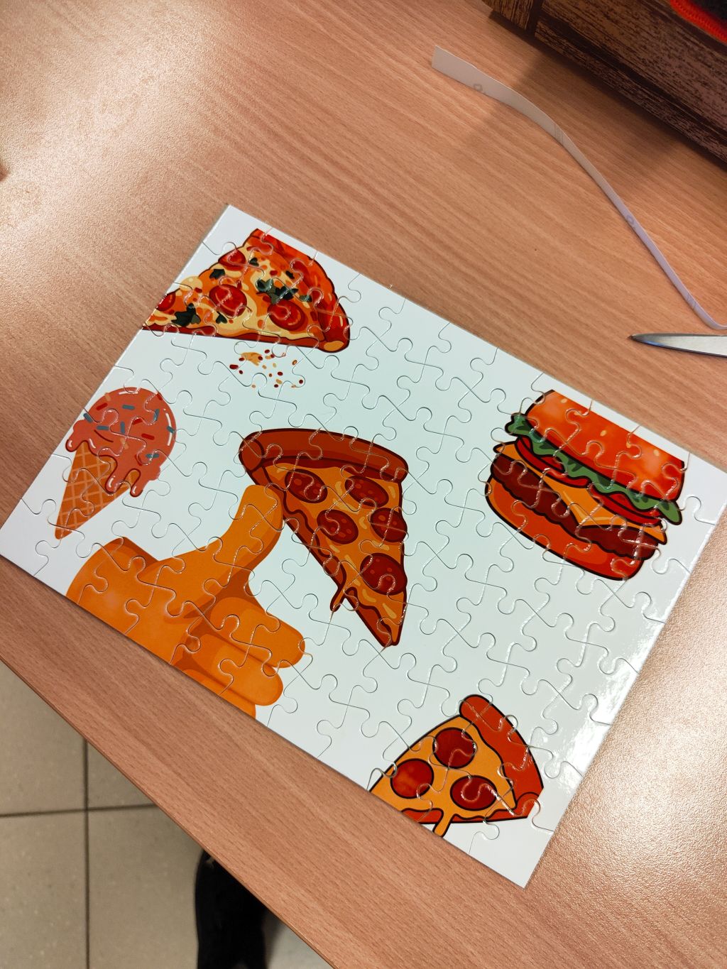 Ułożone na drewnianym blacie puzzle z kolorową grafiką przedstawiającą różne potrawy: trzy kawałki pizzy z pepperoni, burgera oraz loda w rożku.