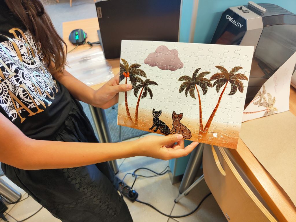 Osoba trzyma w rękach ułożone puzzle przedstawiające dwie postacie kotów na tle palm i chmury.