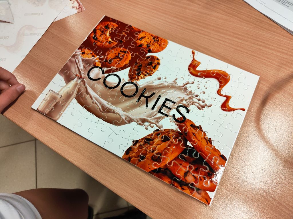 Ułożone na stole puzzle z grafiką przedstawiającą ciasteczka z kawałkami czekolady, rozlane mleko oraz napis „COOKIES”.