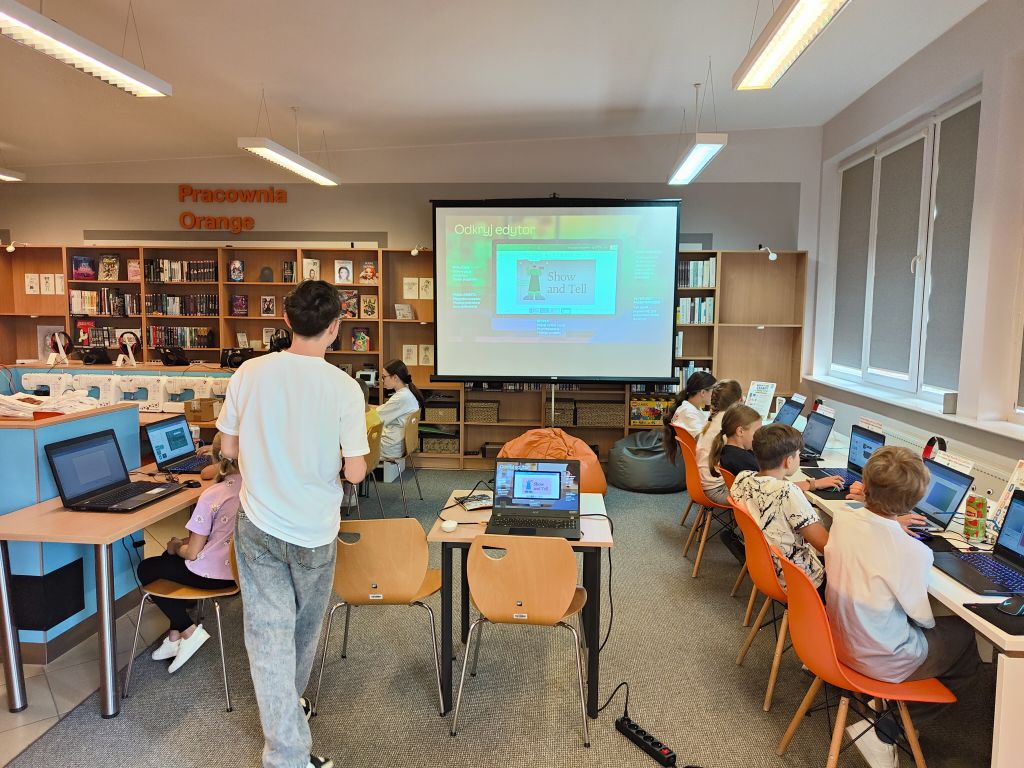 Sala komputerowa w bibliotece, w której dzieci pracują na laptopach przy stołach, z dużym ekranem projekcyjnym z przodu i regałami z książkami w tle.