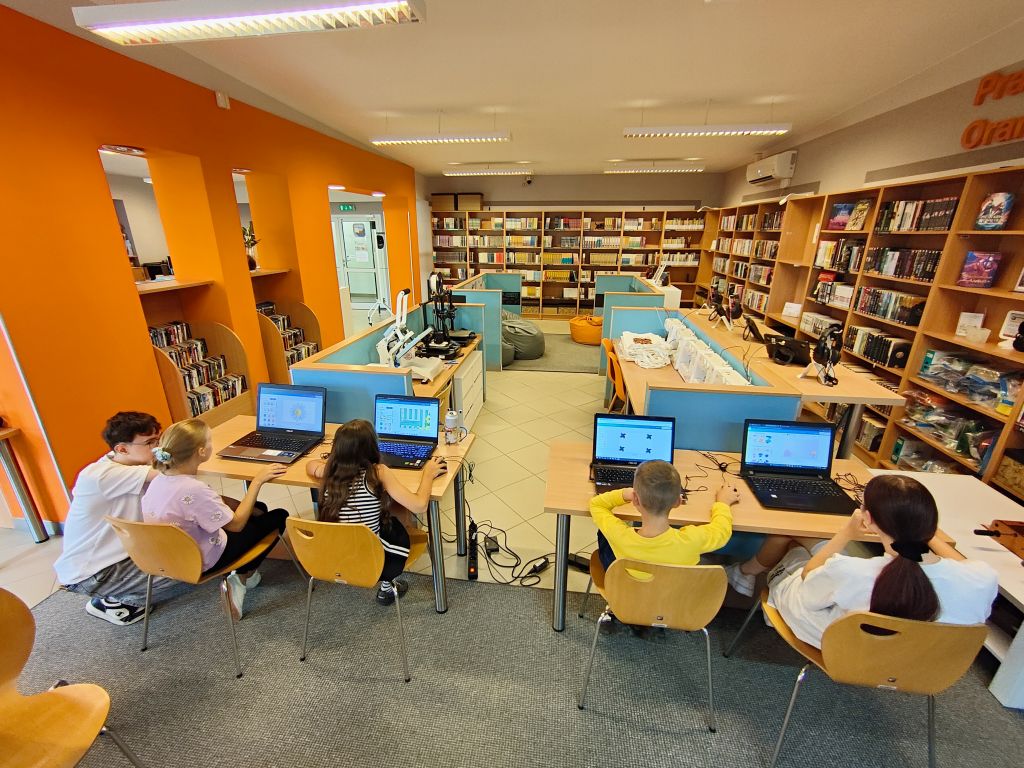 Szerokie ujęcie nowoczesnej sali komputerowej w bibliotece z pomarańczowymi ścianami, regałami na książki i dziećmi siedzącymi przy laptopach.
