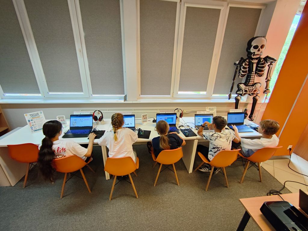 Pięcioro dzieci siedzi tyłem do kadru na pomarańczowych krzesłach przy długim białym stole i pracuje na laptopach pod oknami, obok stojącej dekoracji przedstawiającej szkielet.