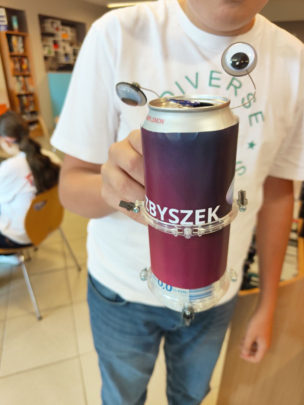 Osoba w białej koszulce trzyma w dłoni robota zrobionego z ciemno-fioletowej puszki z napisem „ZBYSZEK” oraz doklejonymi ruchomymi oczami.