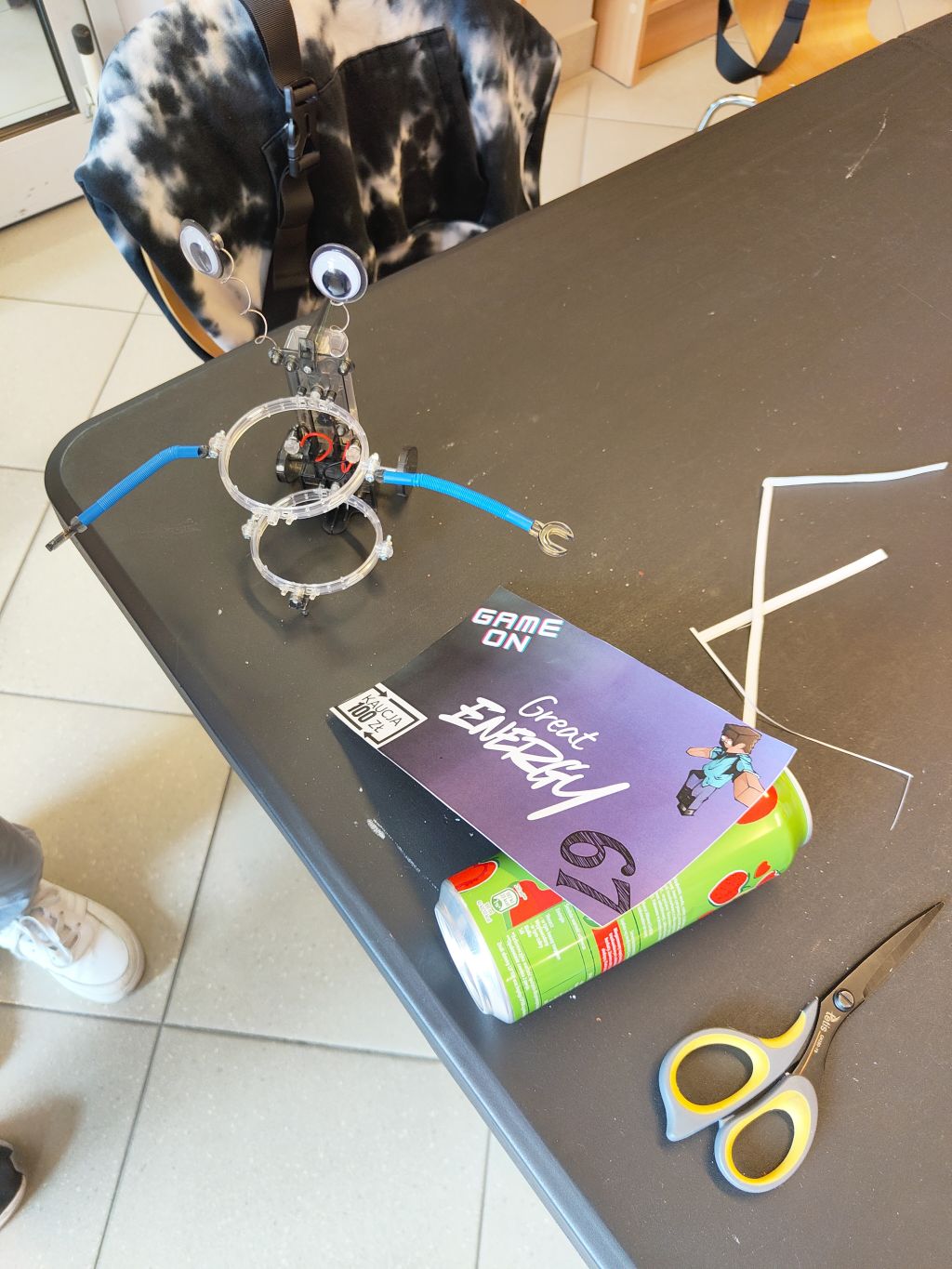 Złożony przezroczysty stelaż robota stojący na stole obok zielonej puszki, kartonowej etykiety i nożyczek.