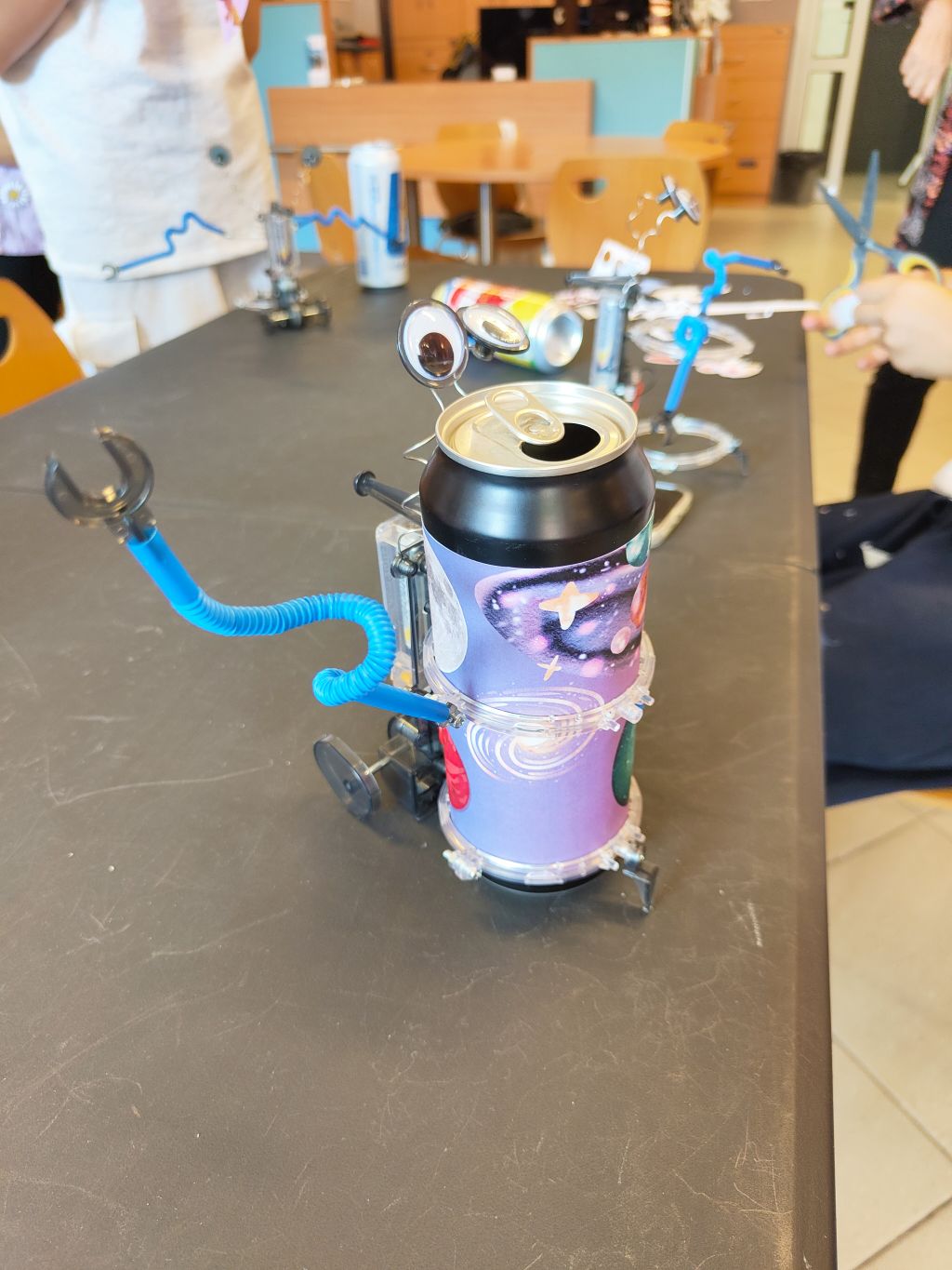 Gotowy robot zbudowany z czarno-fioletowej puszki z kosmicznym wzorem, wyposażony w plastikowe oczy oraz zagięte, niebieskie ramiona.
