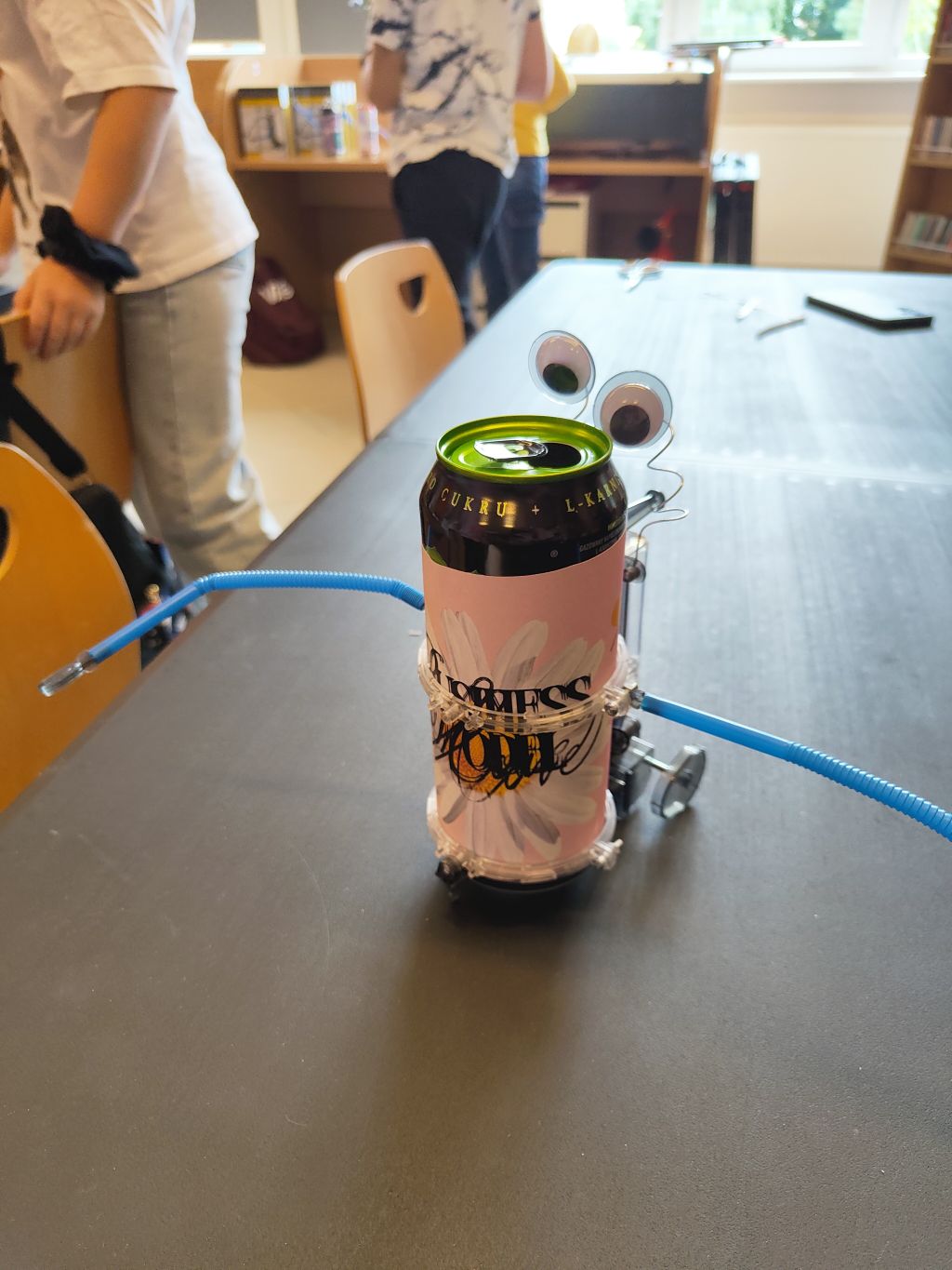 Robot zrobiony z czarnej puszki z różowo-białą etykietą w kwiaty, z doklejonymi wielkimi oczami i niebieskimi słomkami jako ramionami, stojący na stole.