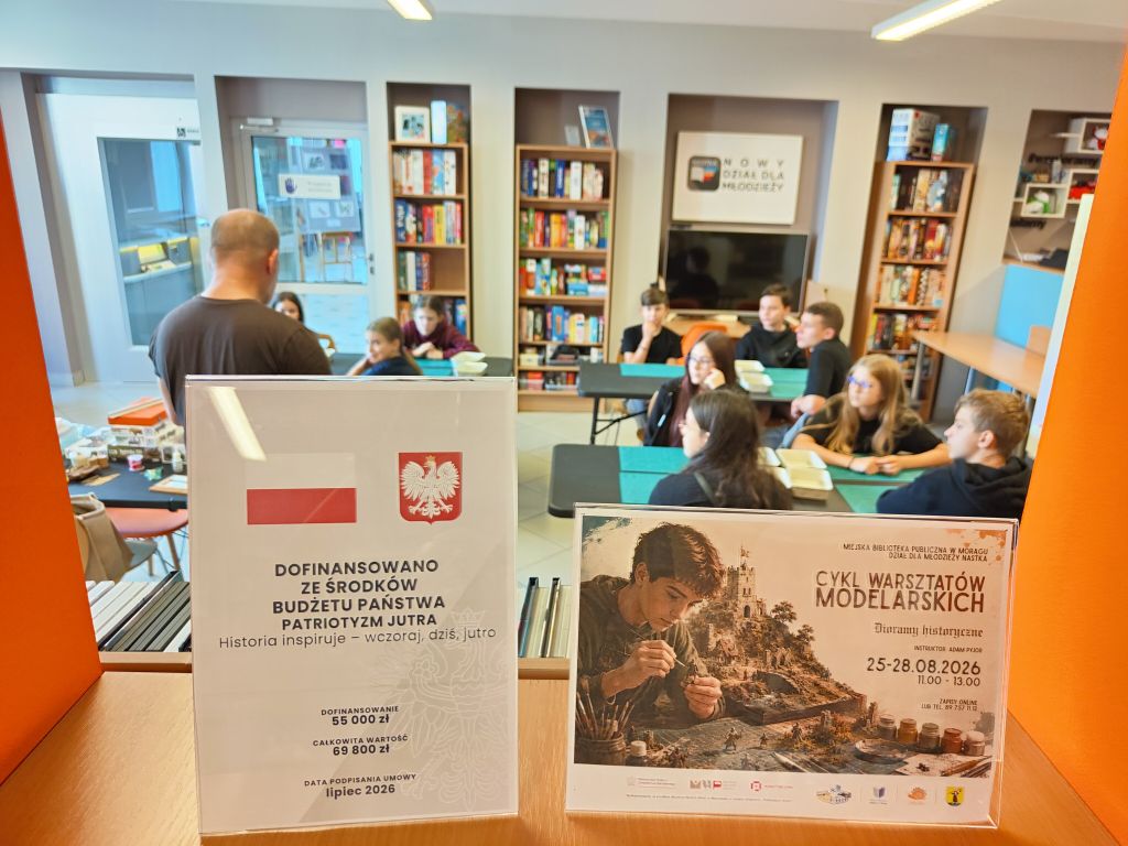 Dwie tabliczki informacyjne dotyczące finansowania i cyklu warsztatów modelarskich, na tle uczestników w sali.