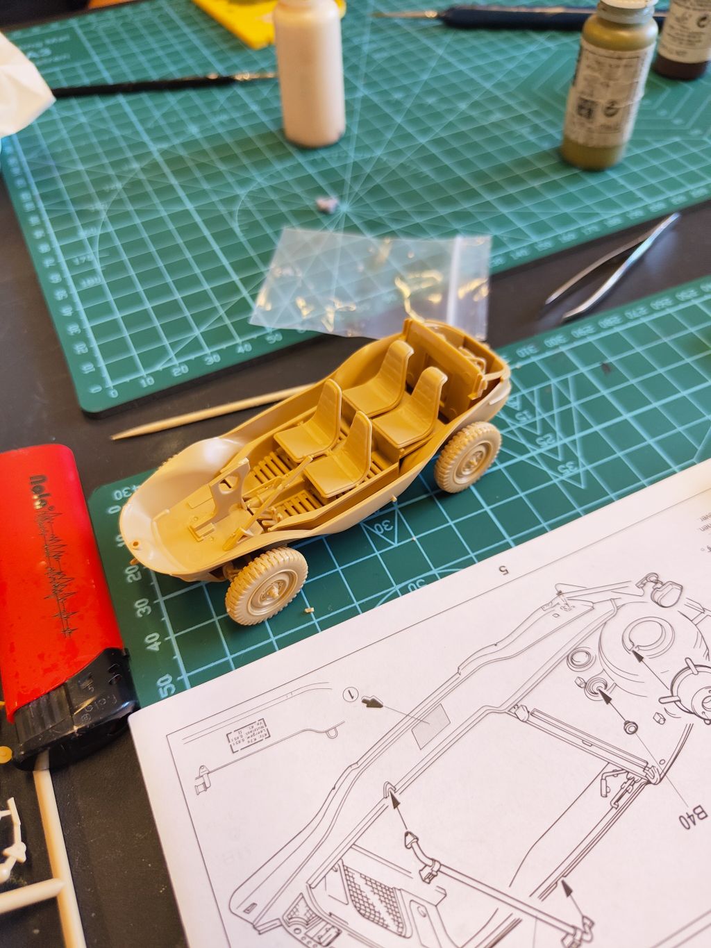 Zbliżenie na częściowo złożony, jasnobrązowy model plastikowego pojazdu wojskowego (typu Schwimmwagen) stojący na zielonej macie obok instrukcji.