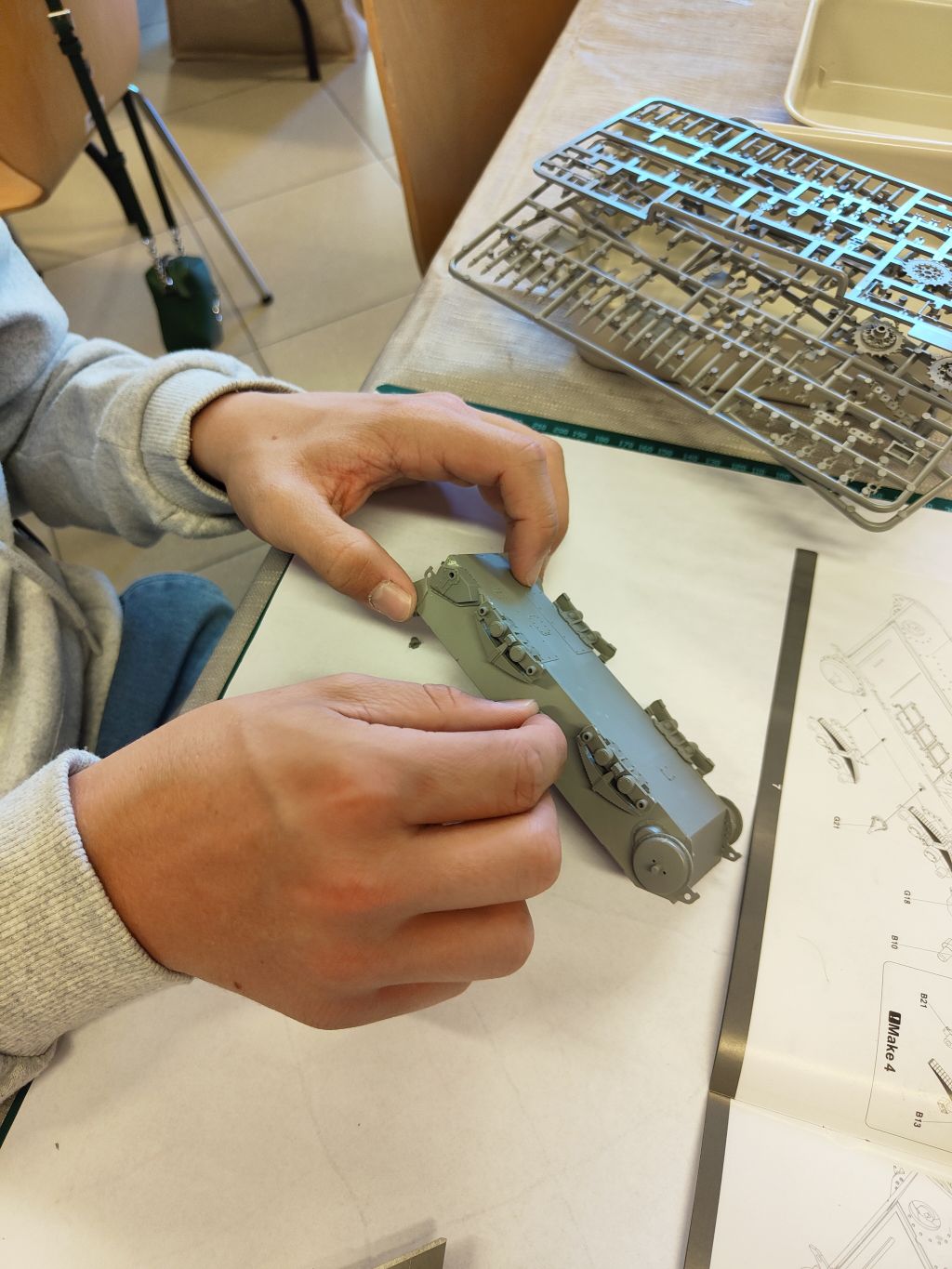 Zbliżenie na dłonie osoby w szarej bluzie, składającej szary plastikowy kadłub modelu czołgu. Obok leżą wypraski z częściami modelu i instrukcja montażu na macie do cięcia.