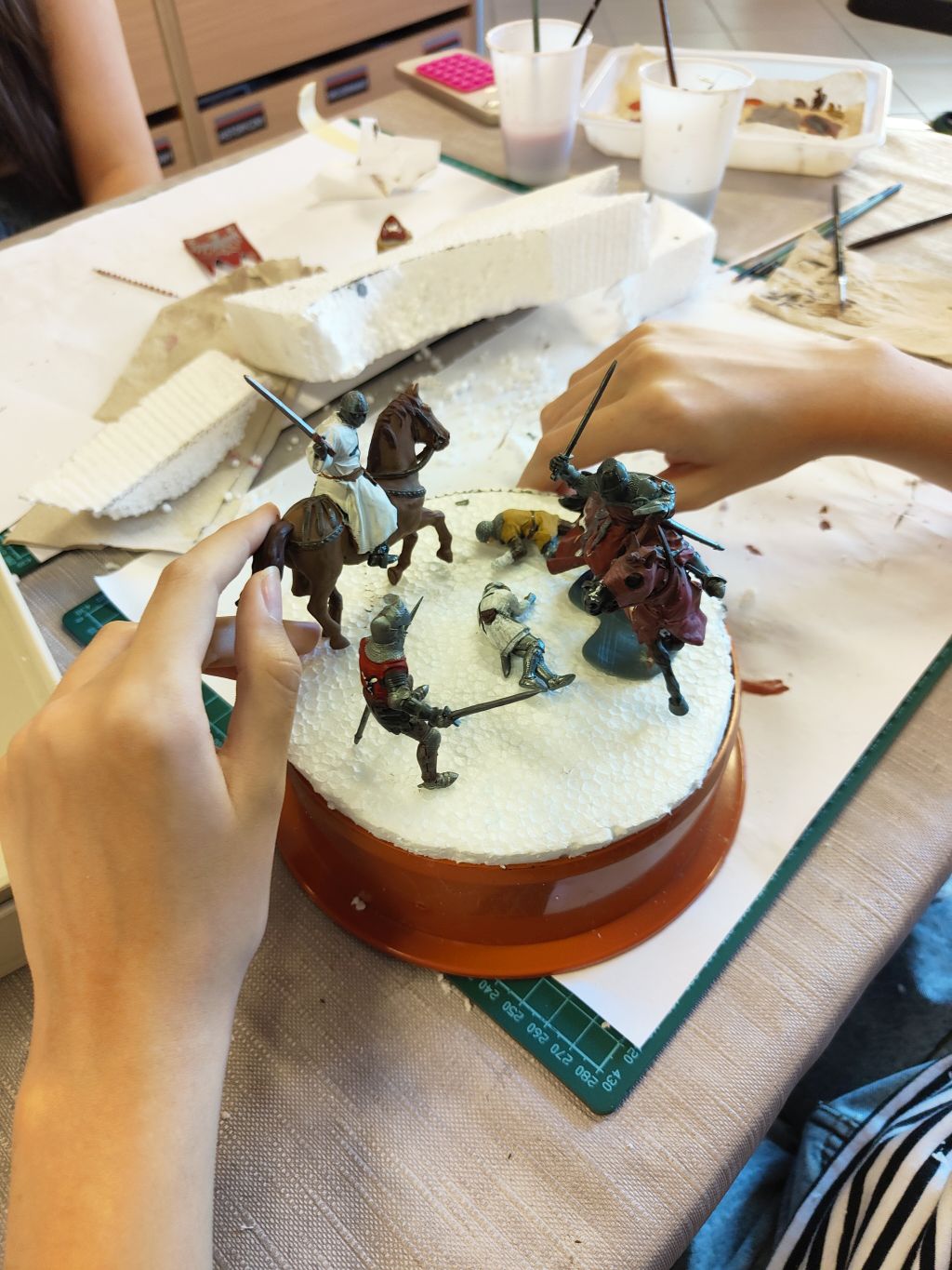 Układanie miniaturowych figurek rycerzy i koni na okrągłej podstawie pokrytej styropianem podczas tworzenia makiet pojedynku.