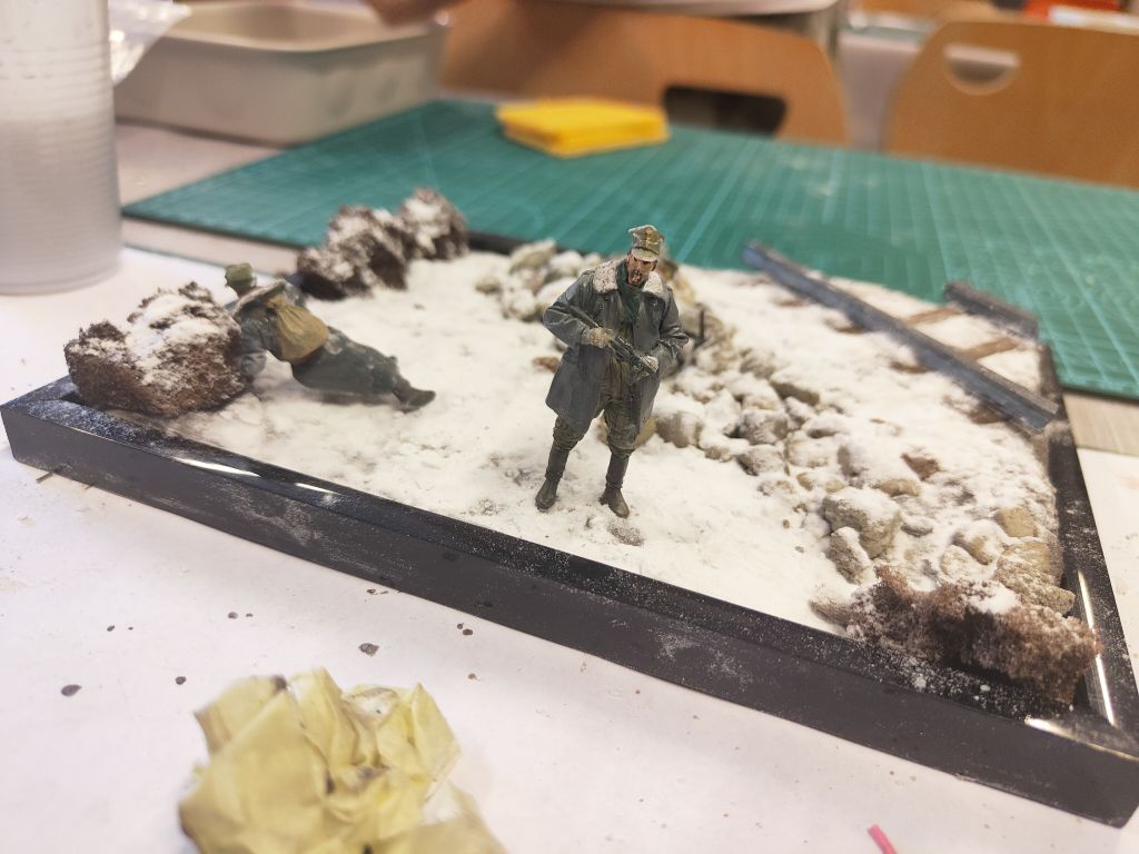 Zbliżenie na fragment makiety, na którym widać modele koni i żołnierzy na trawiastym terenie. Na trawie leży również flaga z godłem Polski.