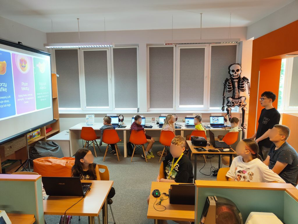 Dzieci i nastolatki pracują przy komputerach i laptopach w jasnej sali warsztatowej. Obok ekranu projekcyjnego stoi model szkieletu.
