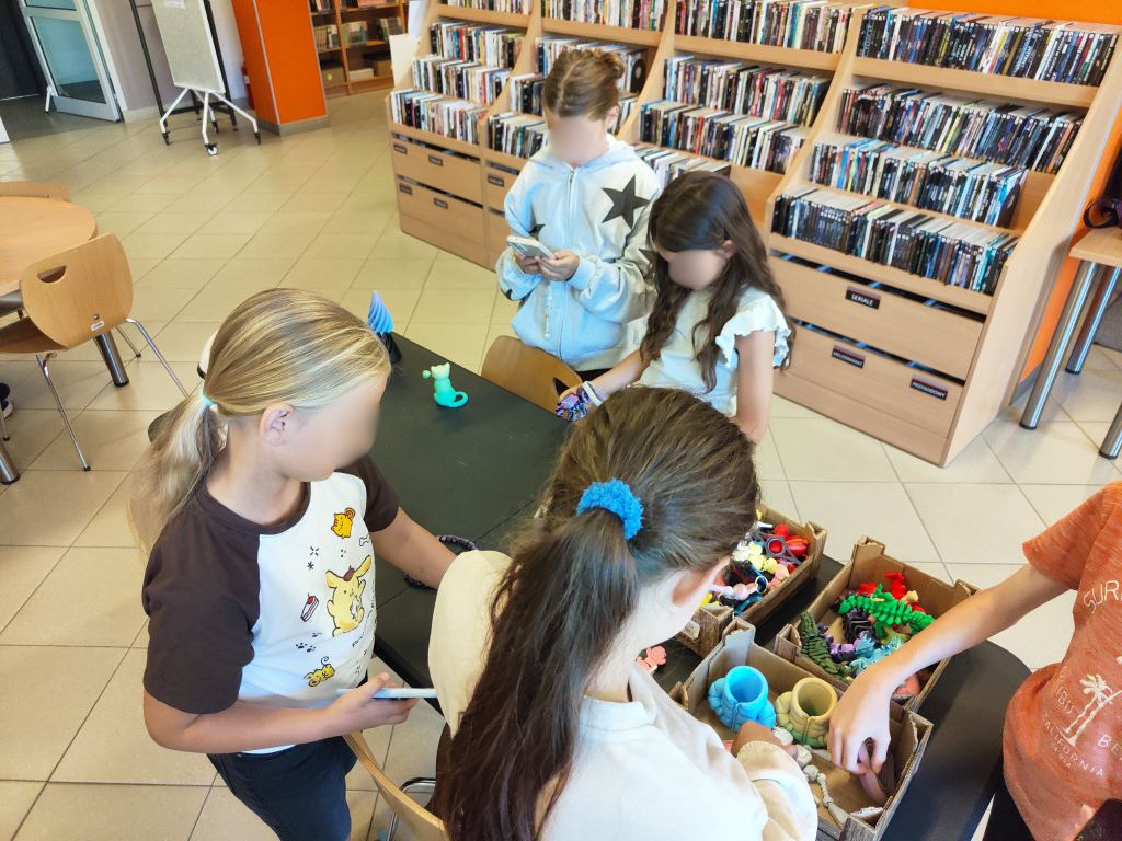 Widok z góry na dzieci stojące przy stole, przeglądające kolorowe elementy wydrukowane na drukarce 3D. W tle stoją regały pełne książek i filmów.