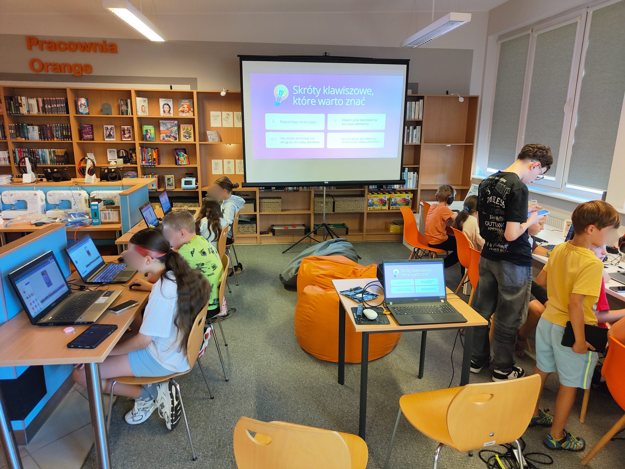Dzieci pracujące na laptopach przy biurkach. Na ekranie projekcyjnym wyświetla się slajd pt. „Skróty klawiszowe, które warto znać”.