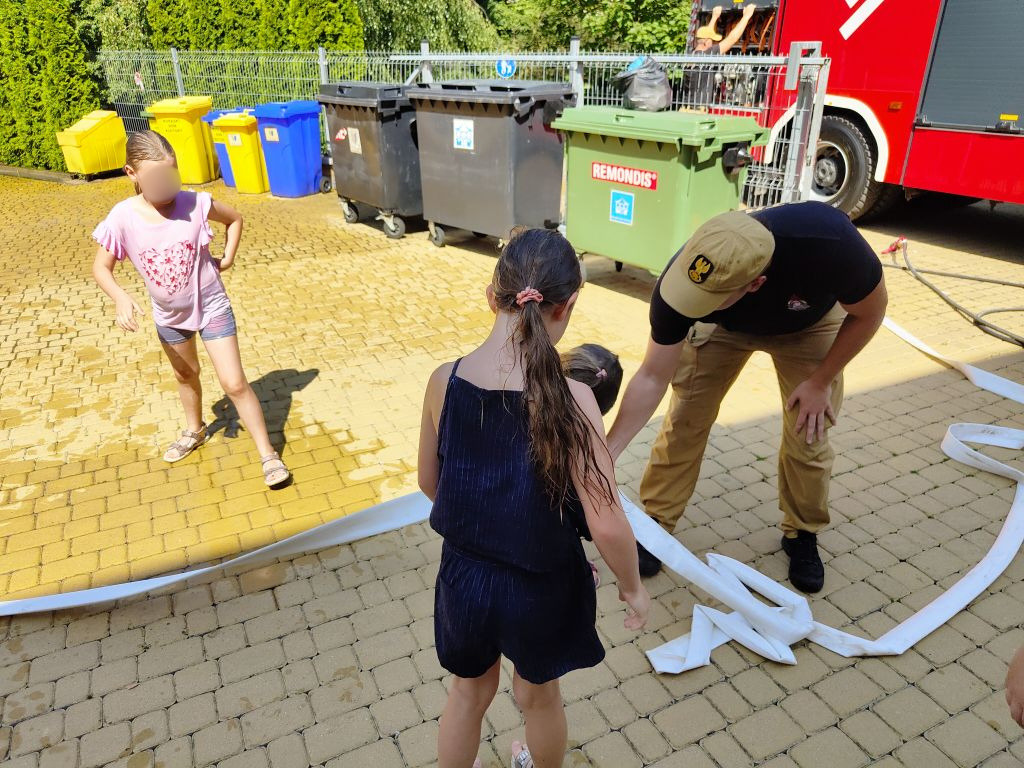 Strażak i dzieci układają na kostce brukowej biały wąż strażacki.