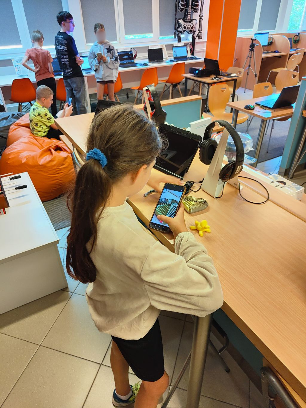 Dziewczynka robia zdjęcie smartfonem złotej ozdobie w kształcie serca, leżącej na blacie obok słuchawek.