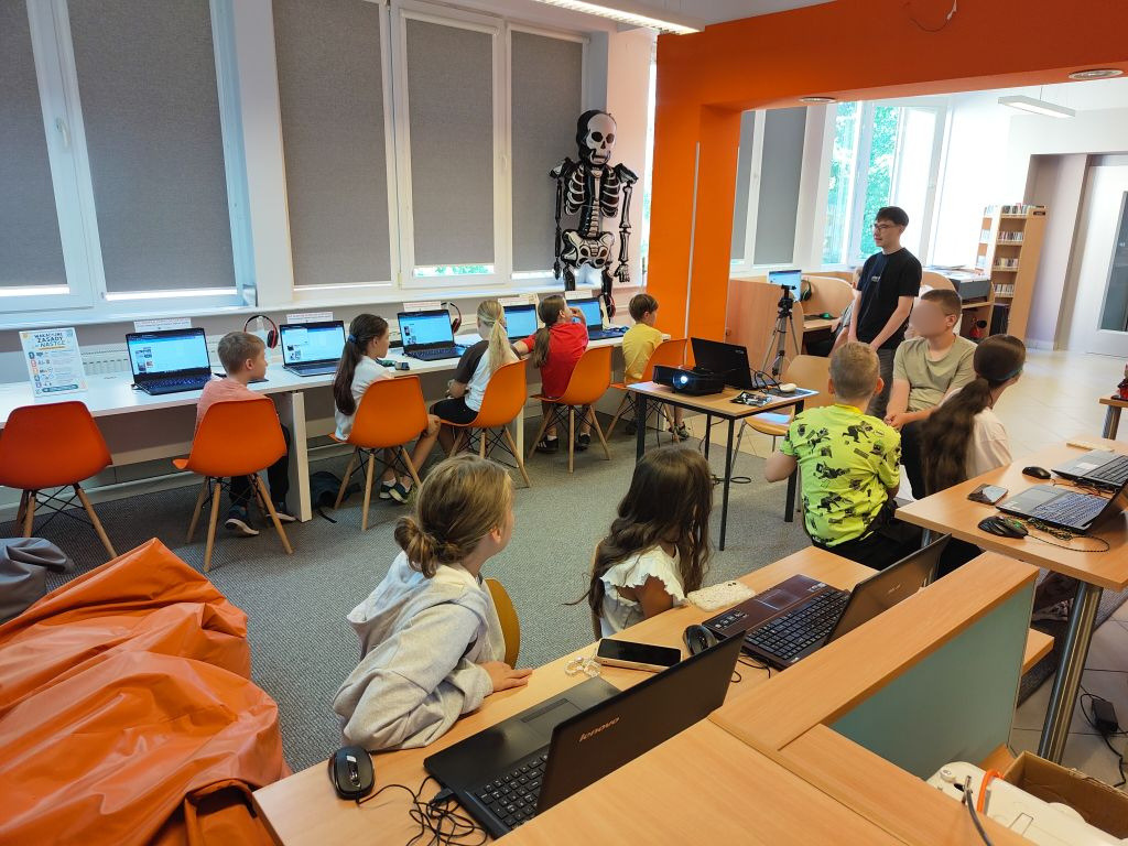 Szerokie ujęcie sali komputerowej – dzieci siedzą przy długim blacie pod oknami z laptopami, podczas gdy na ścianie w tle wisi ozdobny szkielet.