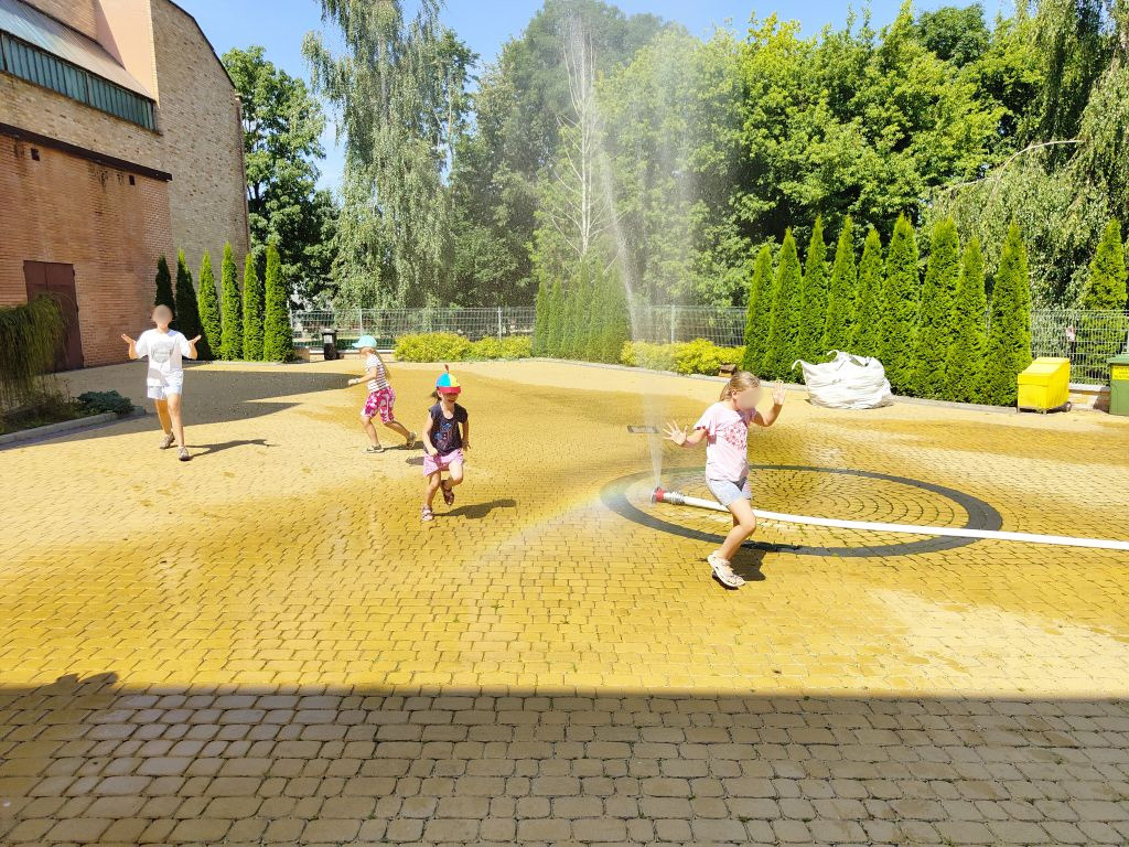 Dzieci bawią się na wybrukowanym placu, biegając wokół wytworzonej z węża kurtyny wodnej.