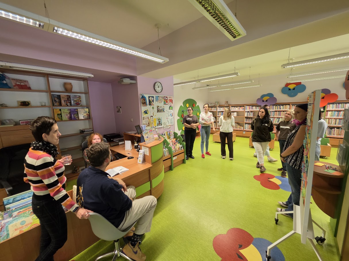 Uczestnicy spotkania sieciującego odwiedzają dział dziecięcy biblioteki i słuchają osoby opowiadającej o jego działalności.