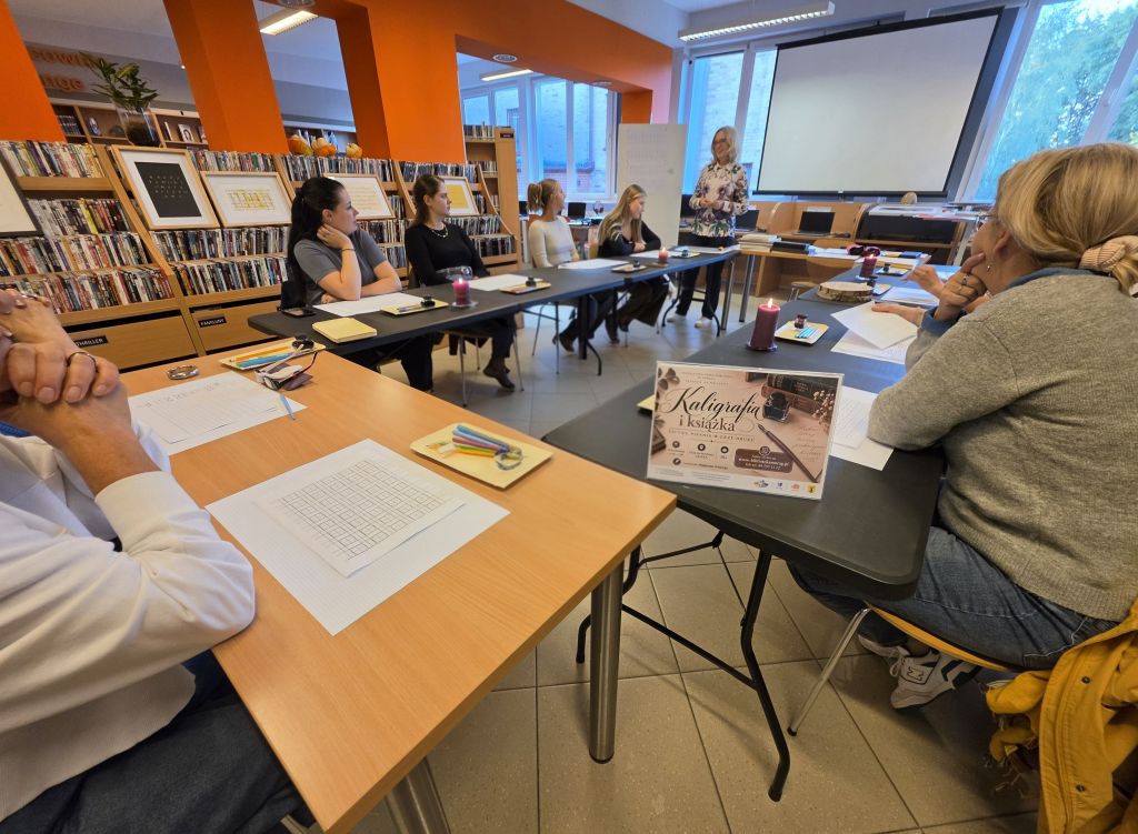 Uczestnicy warsztatów kaligrafii siedzą przy stołach w bibliotece i słuchają prowadzącej stojącej przed ekranem. Na stołach leżą arkusze do ćwiczeń, przybory do pisania i kałamarze.