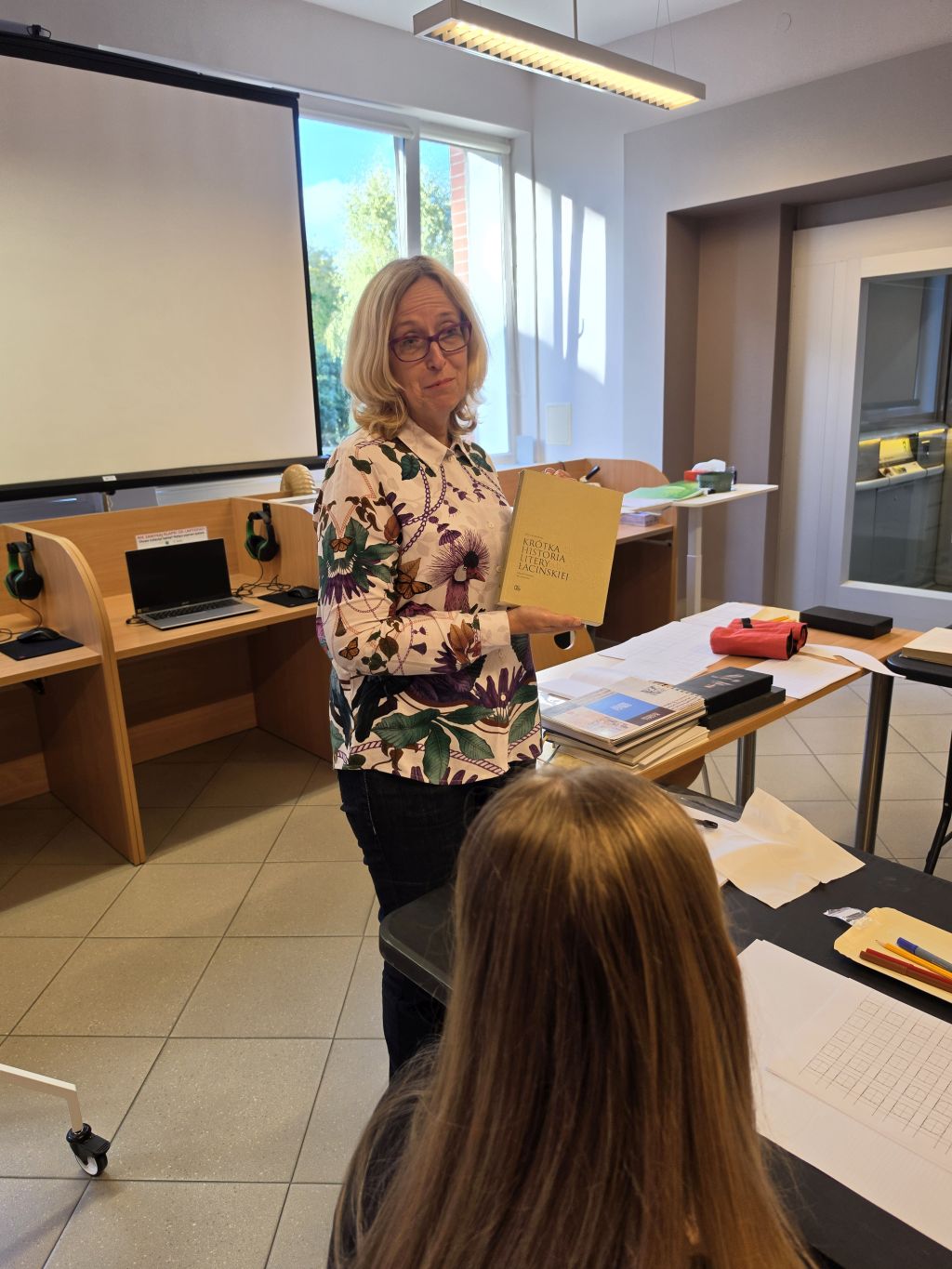 Prowadząca warsztaty prezentuje uczestnikom książkę „Krótka historia litery łacińskiej”. W tle znajduje się ekran projekcyjny i stanowiska komputerowe.