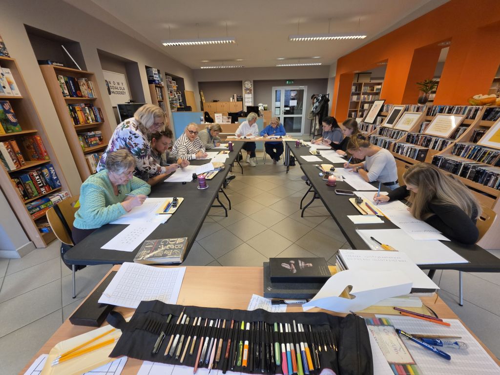 Grupa uczestników wykonuje ćwiczenia kaligraficzne przy stołach w bibliotece. Na pierwszym planie leżą arkusze ćwiczeń oraz zestaw stalówek i pisaków.
