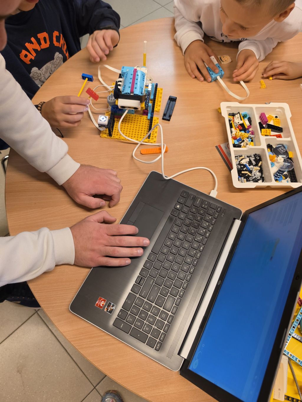 Grupa uczestników pracuje nad robotem LEGO przy stole. Obok konstrukcji znajduje się laptop i pojemnik z elementami zestawu.