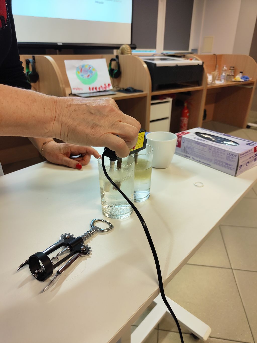Osoba zanurzająca urządzenie elektryczne z czarnym przewodem w szklance z wodą na stole.