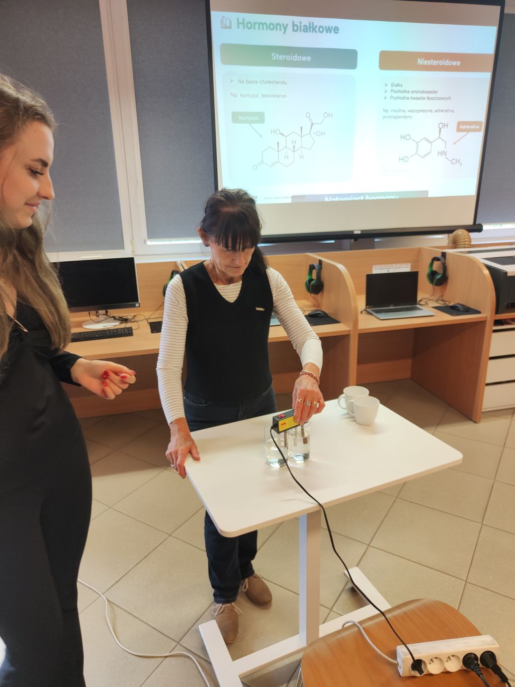 Kobieta w średnim wieku wykonuje eksperyment chemiczny, zanurzając elektrolizer w szklankach z wodą na tle rzutnika z prezentacją o hormonach.