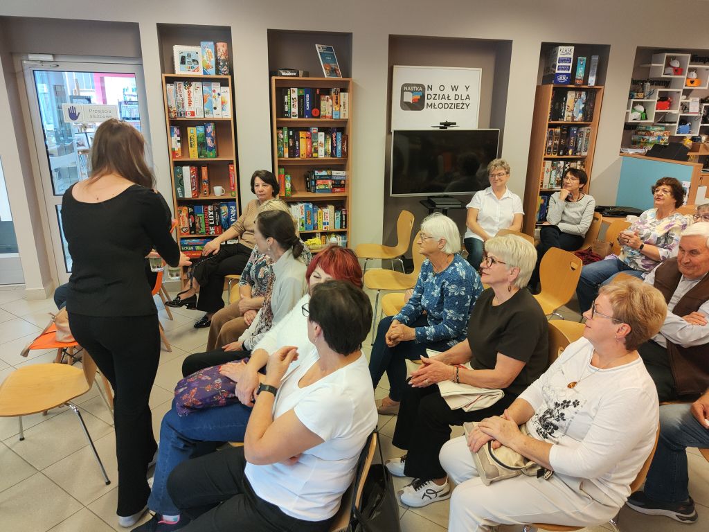 Grupa osób siedzi na krzesłach w sali bibliotecznej, przysłuchując się prelekcji prowadzonej przez stojącą kobietę.