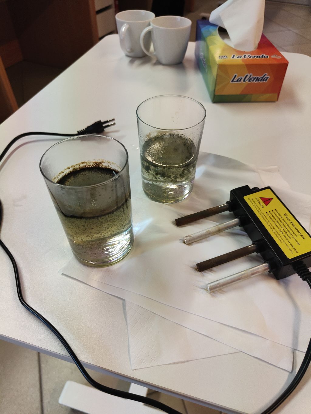 Stół laboratoryjny z dwiema szklankami wody po elektrolizie, elektrolizerem oraz pudełkiem chusteczek.