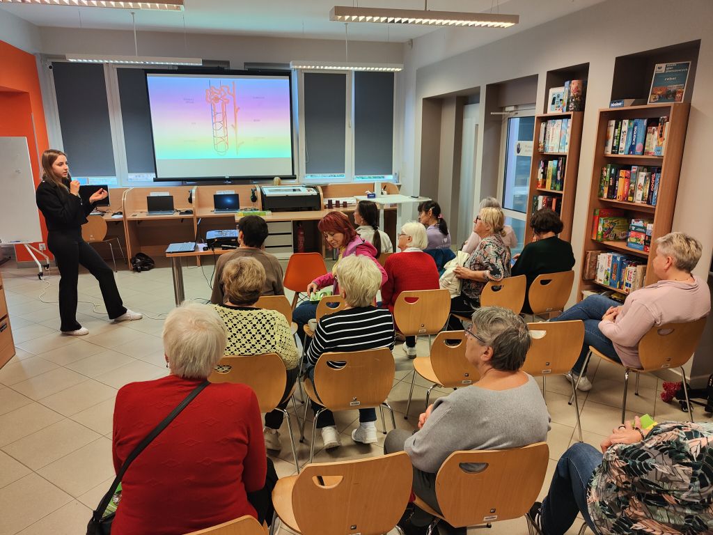 Prelegentka prowadzi prezentację przed grupą słuchaczy siedzących na krzesłach w sali.