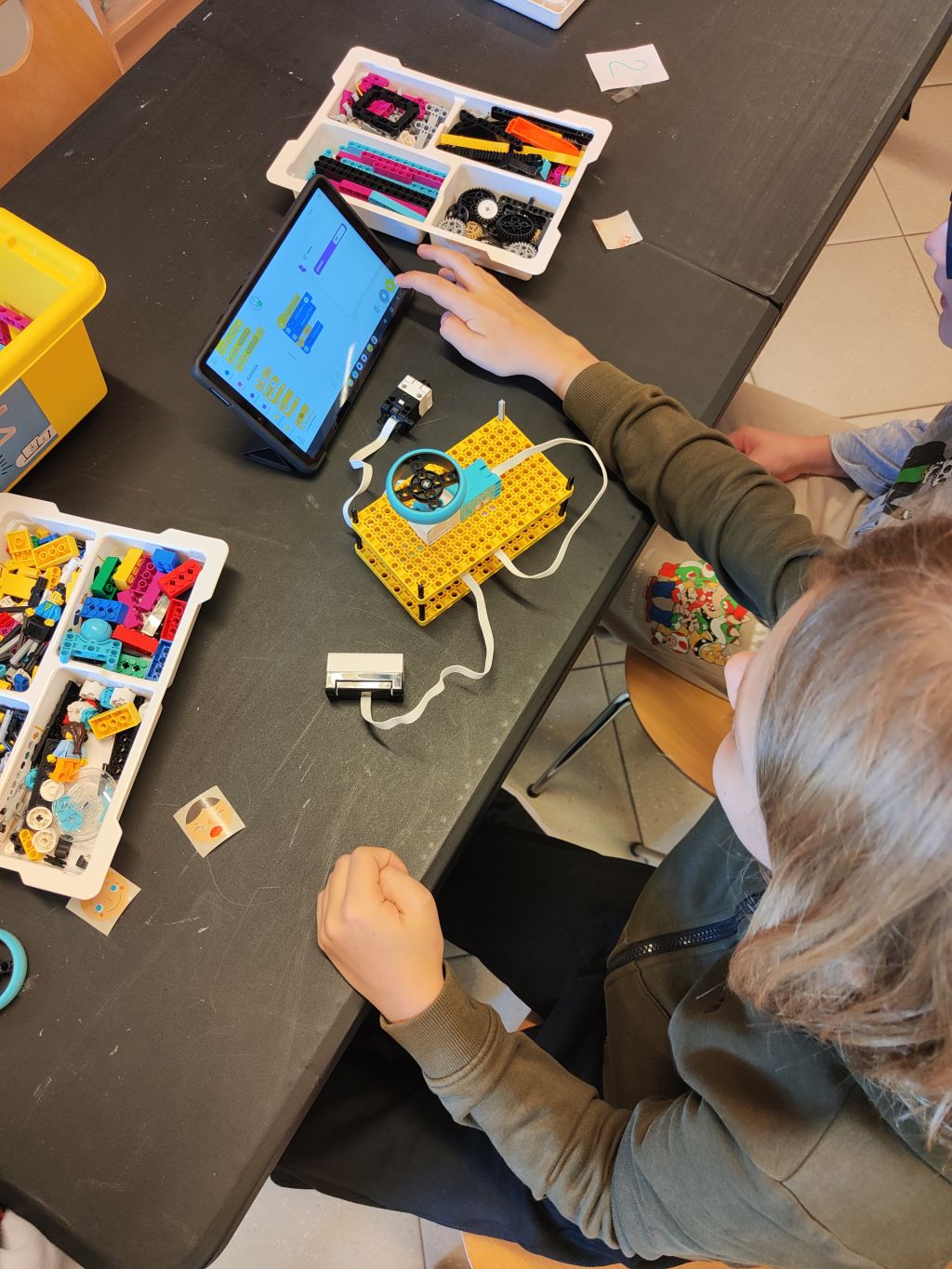 Dwoje uczestników programuje konstrukcję LEGO przy użyciu tabletu. Na stole znajdują się pojemniki z klockami i elementami technicznymi.