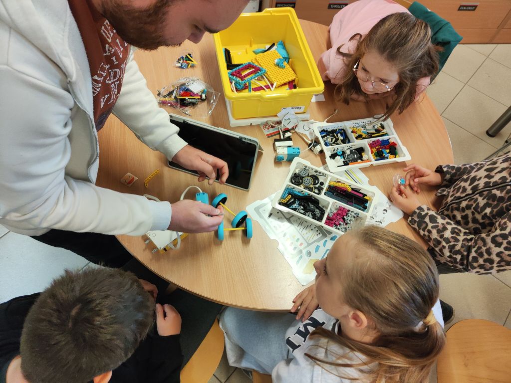 Prowadzący pomaga grupie uczestników przy składaniu robota LEGO. Na stole znajdują się zestawy klocków, tablet i części konstrukcyjne.