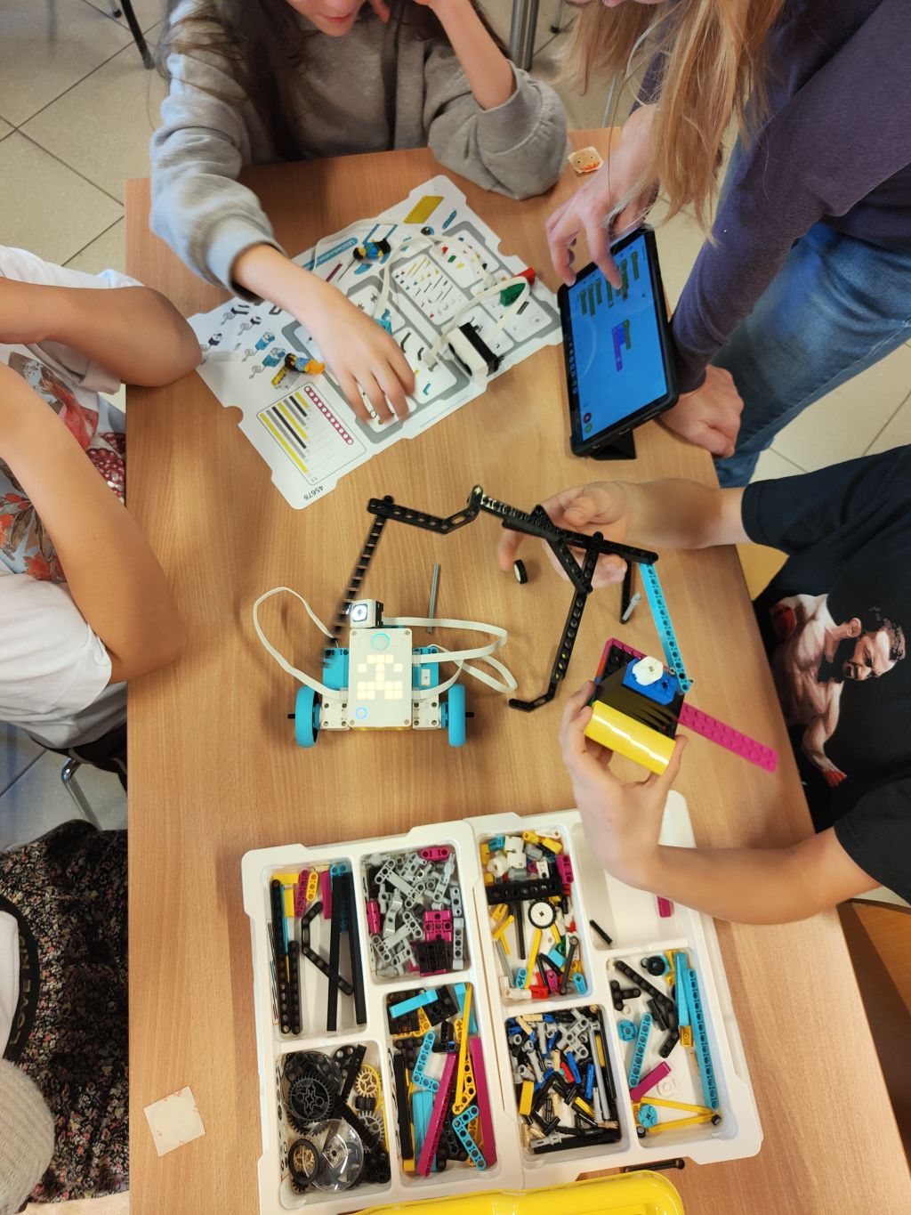 Grupa uczestników wspólnie buduje robota z LEGO i korzysta z tabletu z programem do kodowania. Na stole rozłożone są instrukcje i elementy zestawu.