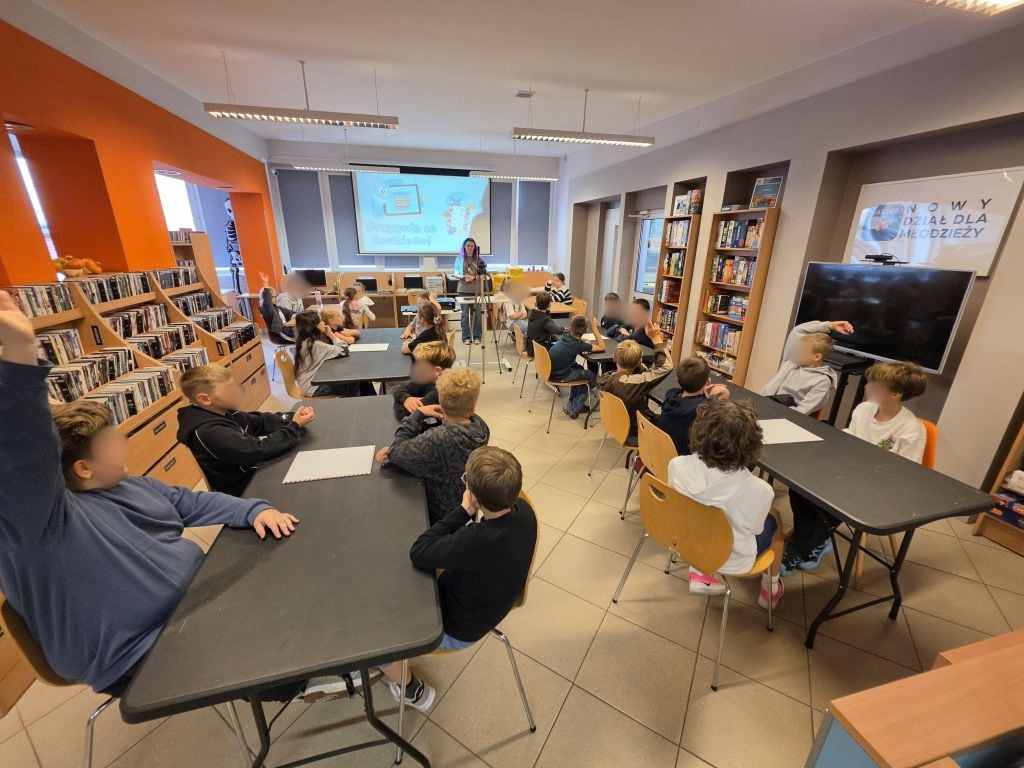 Dzieci siedzące przy stolikach w sali bibliotecznej podczas zajęć.
