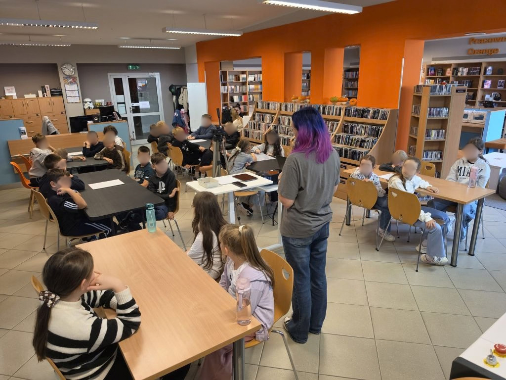 Prowadząca z fioletowymi włosami stojąca przed grupą dzieci siedzących przy stolikach.