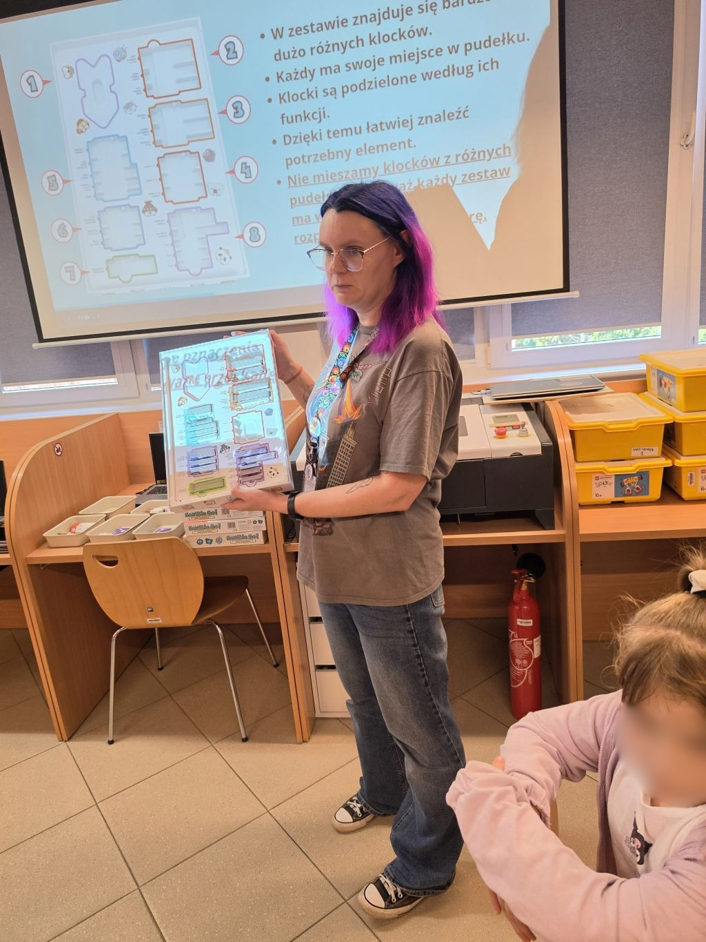 Prowadząca prezentująca instrukcję z klockami edukacyjnymi na tle ekranu multimedialnego.