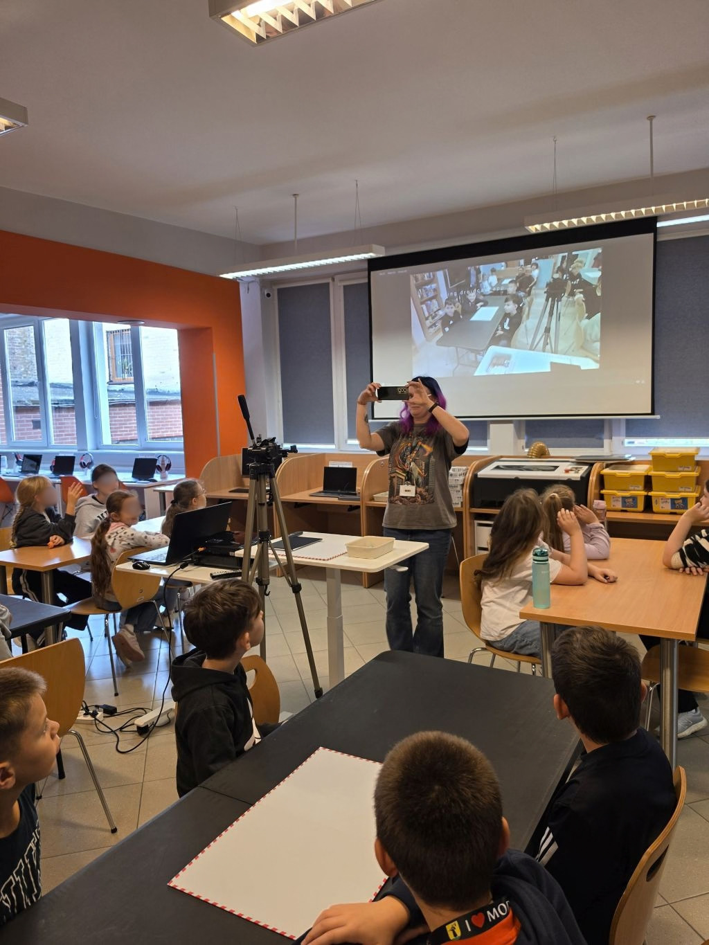Prowadząca nagrywająca tabletem stanowisko z matą edukacyjną, widoczni uczestnicy zajęć.