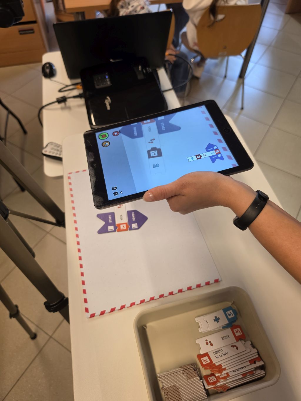 Tablet z programem blokowym trzymany nad matą z kodowaniem i zestawem klocków.