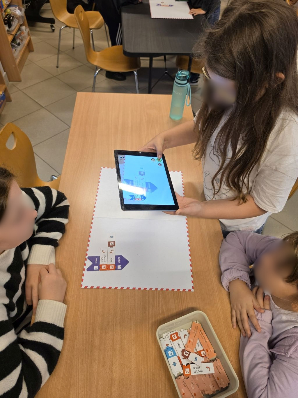 Dziewczynka trzymająca tablet nad matą z ułożonymi klockami kodującymi, obok siedzą inni uczestnicy.