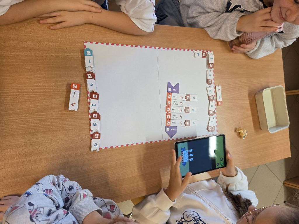 Dzieci przy stoliku układające sekwencję kodowania na białej macie, trzymając tablet.