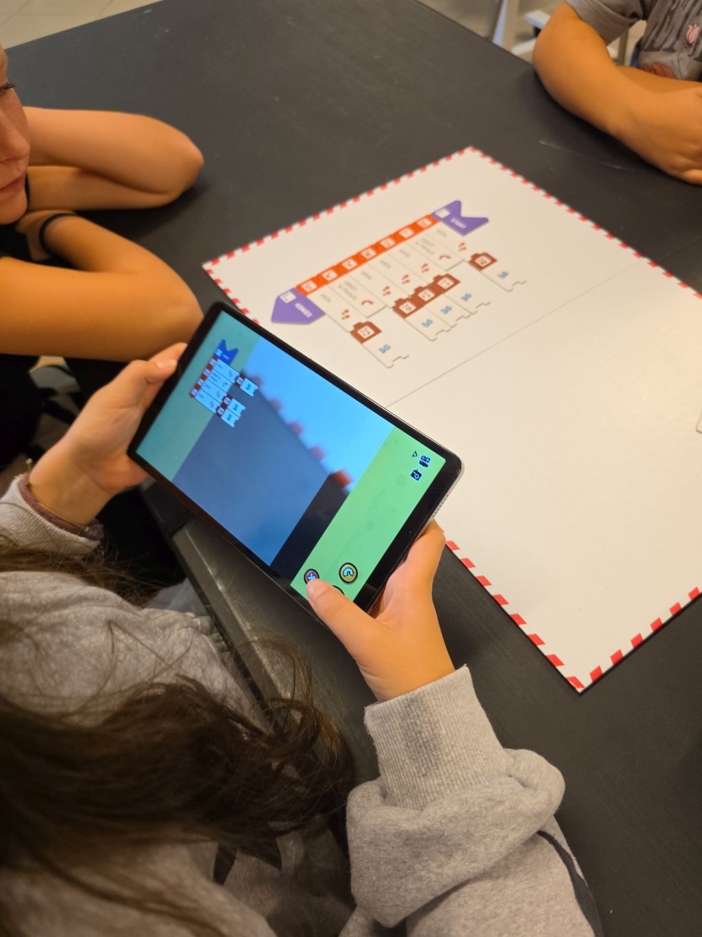 Dziewczynka trzymająca tablet z widokiem aplikacji edukacyjnej nad matą z kodem.