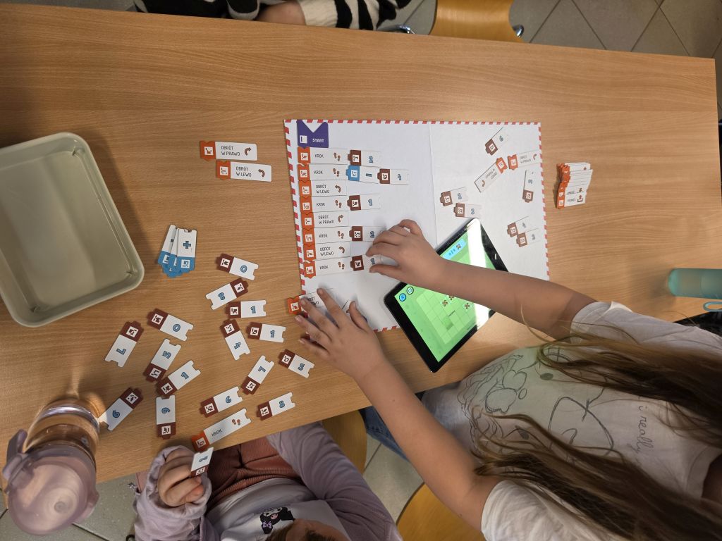 Dzieci pracujące z matą edukacyjną, klockami i tabletem na drewnianym stoliku.