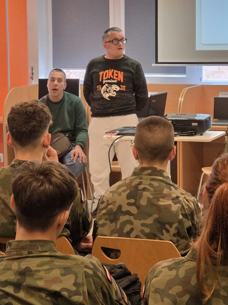 Uczniowie w mundurach wojskowych siedzą na krzesłach i słuchają prowadzących. W tle widoczne są regały z książkami i ekran z prezentacją.